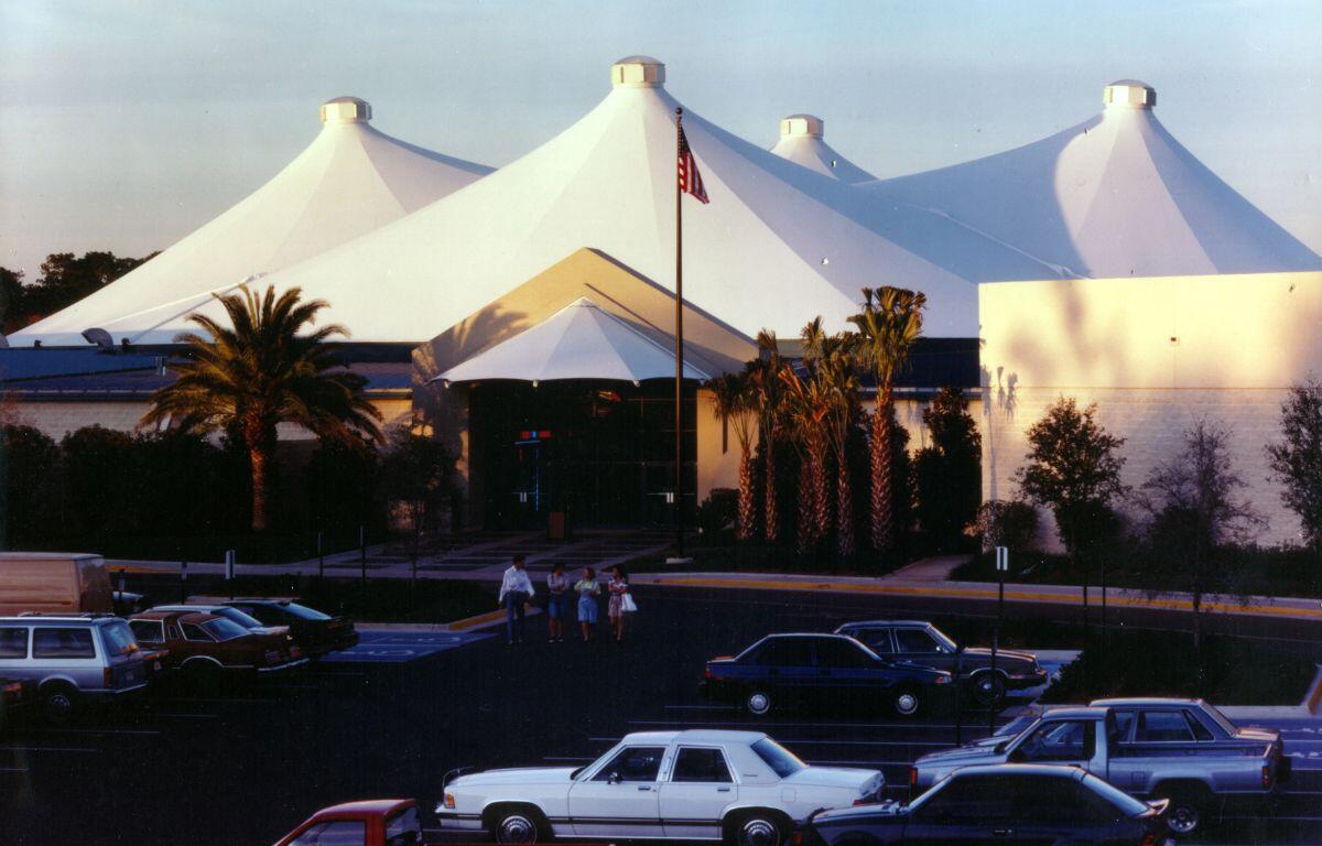 An enclosed mall with a white tent canopy covering and brick walls. Palm trees decorate the entrance with antique cars in the parking lot.