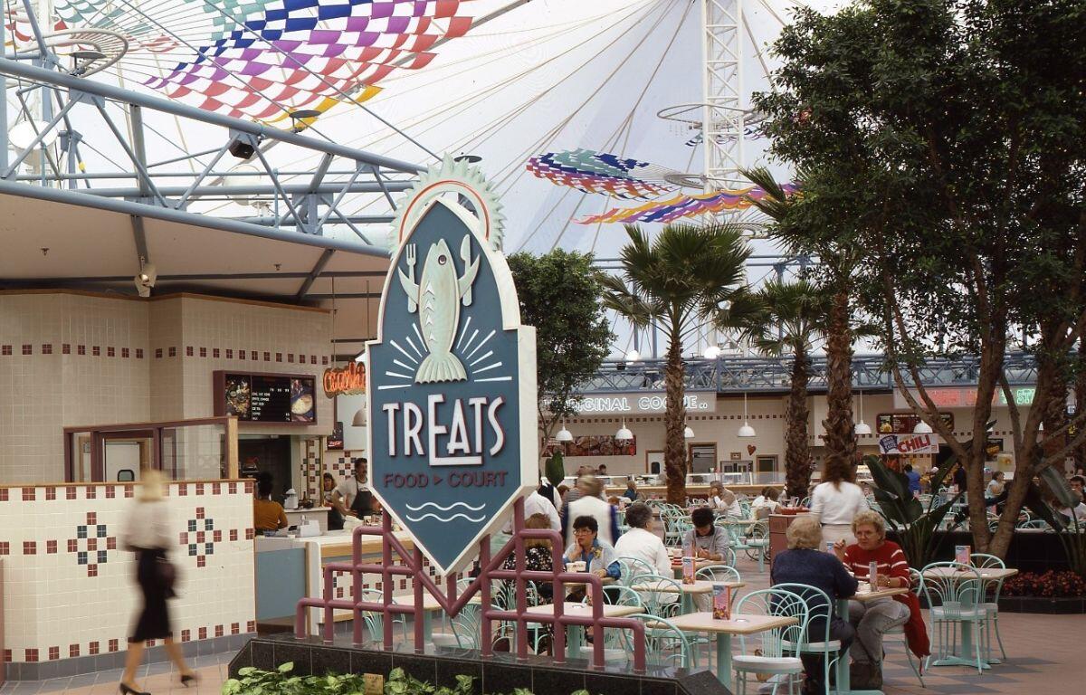 A sign that says, "TrEats," in the foreground. Behind the sign are decorative palm trees and colorful flags suspended underneath a white canopy. People sit at tables and booth dining.