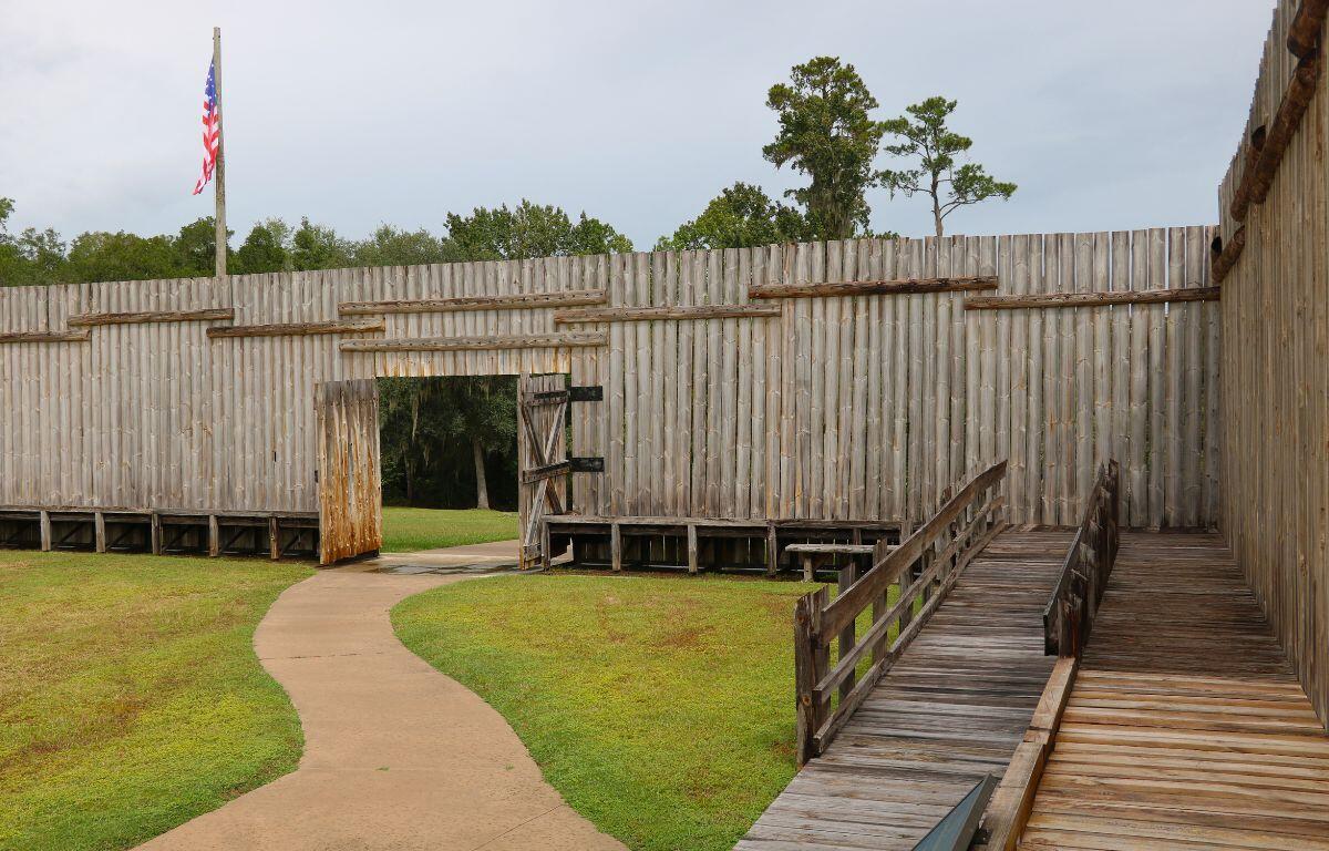 Wooden walls have an open door that leads to an open field and dense forest. A flagpole with an American flag stands beyond the wall and a wooden ramp leads to a thin platform at the corner of the wall. A small path leads through the door and into the area.