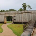 Wooden walls have an open door that leads to an open field and dense forest. A flagpole with an American flag stands beyond the wall and a wooden ramp leads to a thin platform at the corner of the wall. A small path leads through the door and into the area.