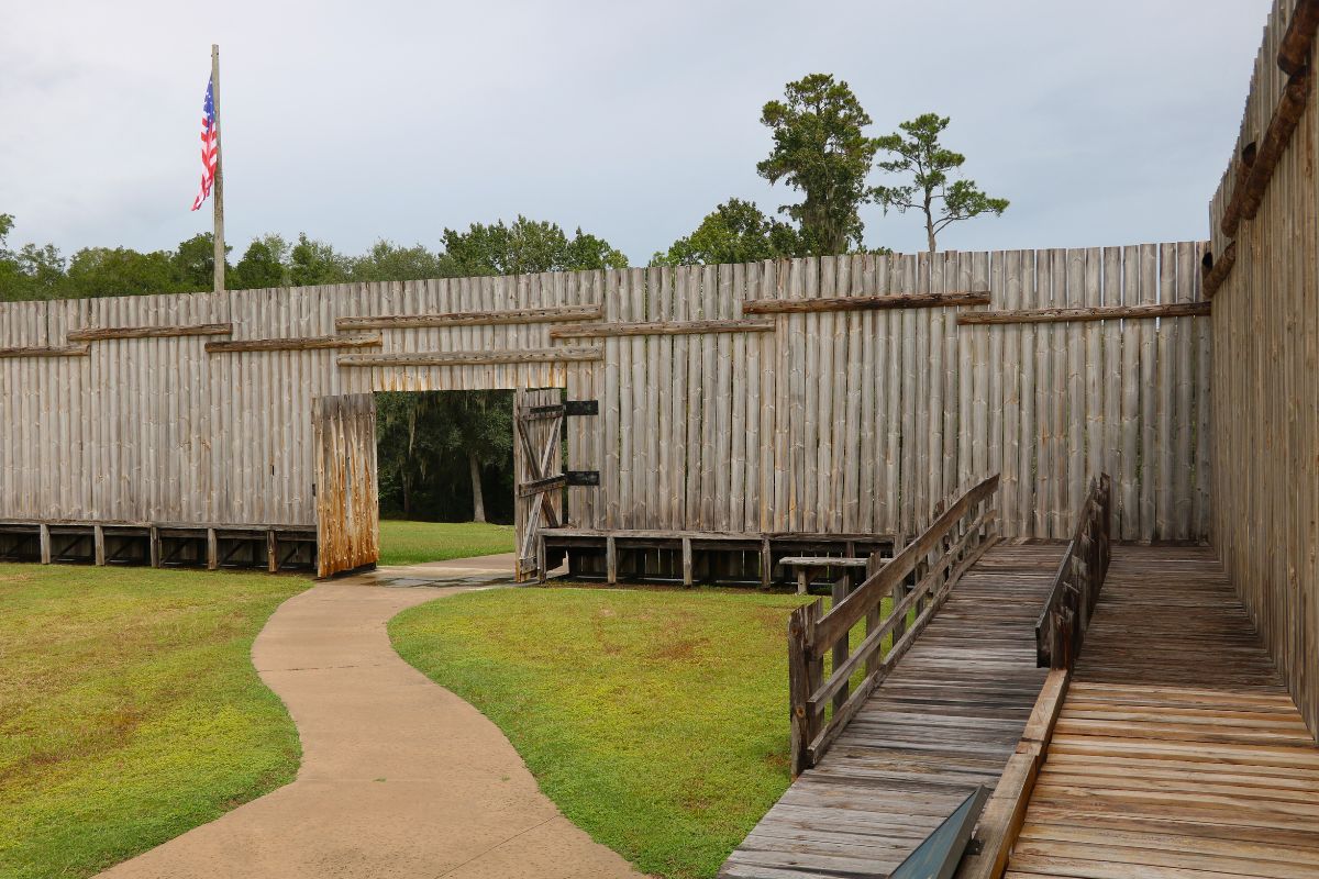 Wooden walls have an open door that leads to an open field and dense forest. A flagpole with an American flag stands beyond the wall and a wooden ramp leads to a thin platform at the corner of the wall. A small path leads through the door and into the area.