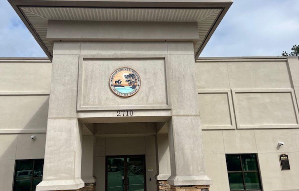The facade of a building, with a off beige trim hanging over the entrance, off beige concrete siding, an insignia handing over the entrance with lettering an image, glass doors in the center background, glass window to the right of a concrete column.