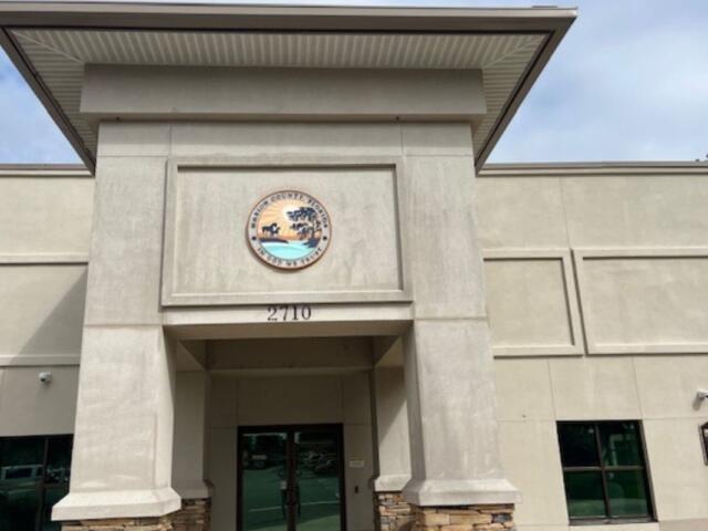The facade of a building, with a off beige trim hanging over the entrance, off beige concrete siding, an insignia handing over the entrance with lettering an image, glass doors in the center background, glass window to the right of a concrete column.