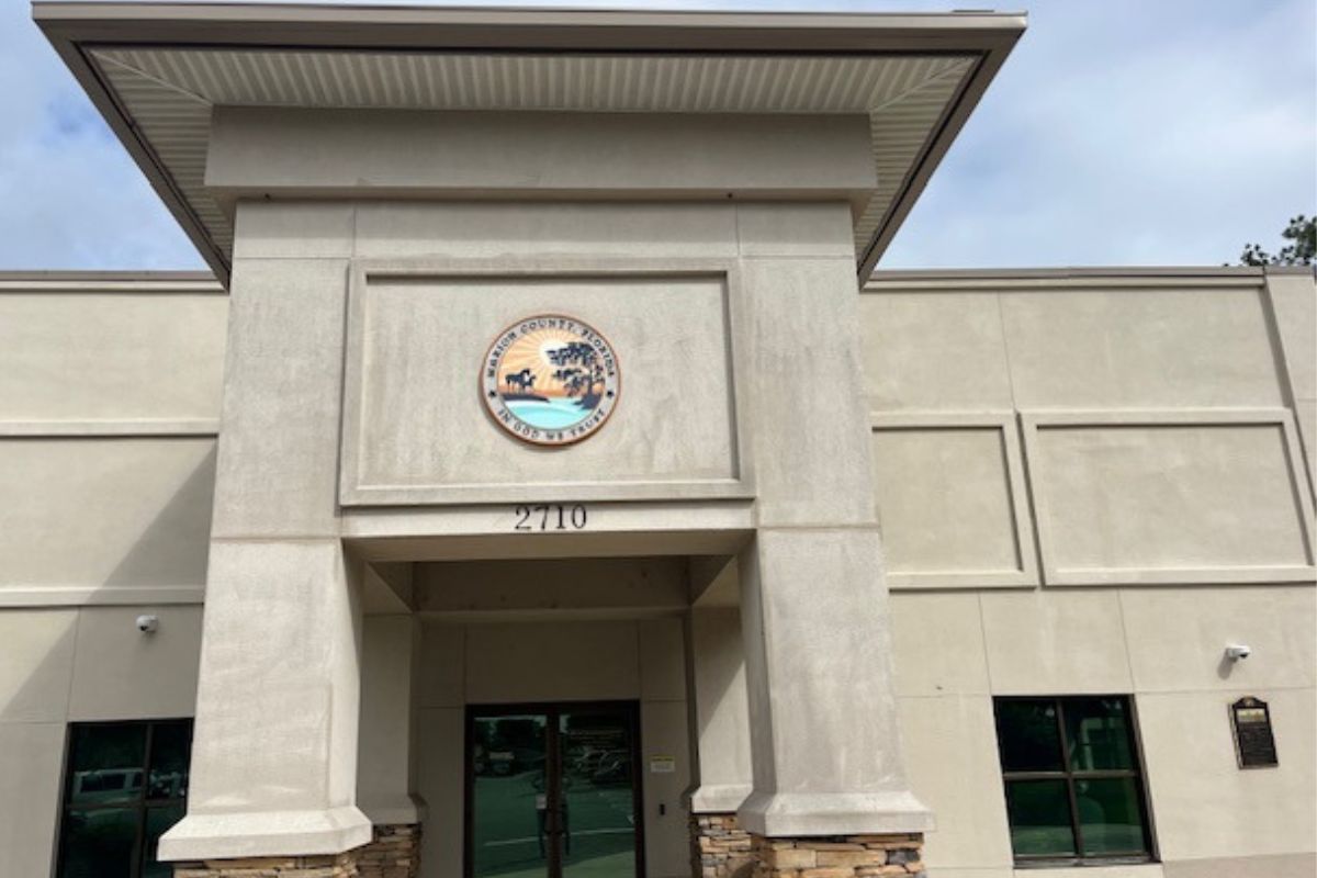The facade of a building, with a off beige trim hanging over the entrance, off beige concrete siding, an insignia handing over the entrance with lettering an image, glass doors in the center background, glass window to the right of a concrete column.
