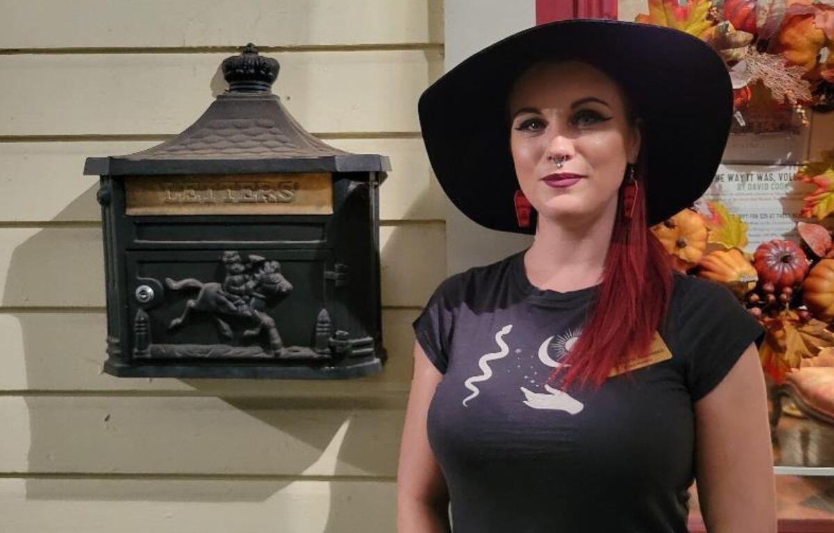 A woman with red hair, a black hat and a black shirt with white graphics stands in front of a beige house with a Halloween themed wreath hanging on the door and a decorated mailbox hanging on the wall with a horse design on it.