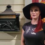 A woman with red hair, a black hat and a black shirt with white graphics stands in front of a beige house with a Halloween themed wreath hanging on the door and a decorated mailbox hanging on the wall with a horse design on it.