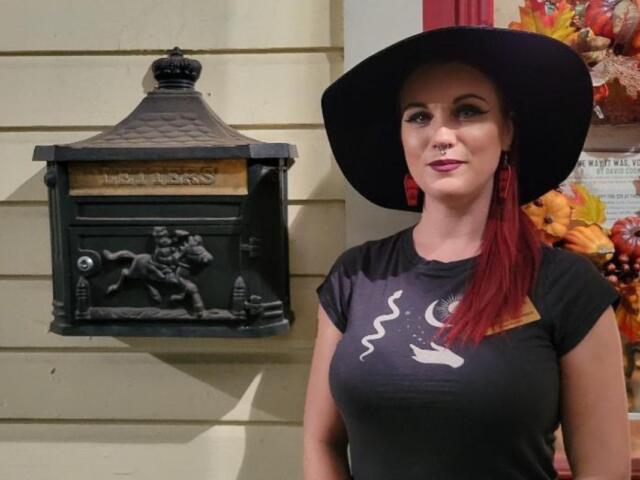 A woman with red hair, a black hat and a black shirt with white graphics stands in front of a beige house with a Halloween themed wreath hanging on the door and a decorated mailbox hanging on the wall with a horse design on it.