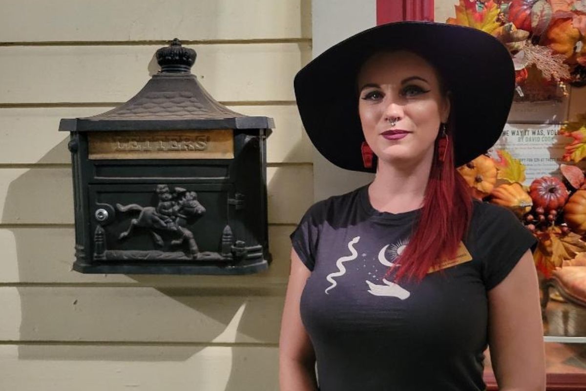 A woman with red hair, a black hat and a black shirt with white graphics stands in front of a beige house with a Halloween themed wreath hanging on the door and a decorated mailbox hanging on the wall with a horse design on it.