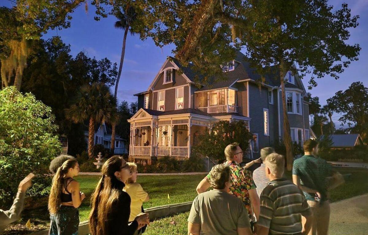 A group of people walk on a sidewalk toward a two-story gray house with white accents and a dark gray roof. Grass and trees surround the property with other houses in the background.