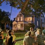 A group of people walk on a sidewalk toward a two-story gray house with white accents and a dark gray roof. Grass and trees surround the property with other houses in the background.