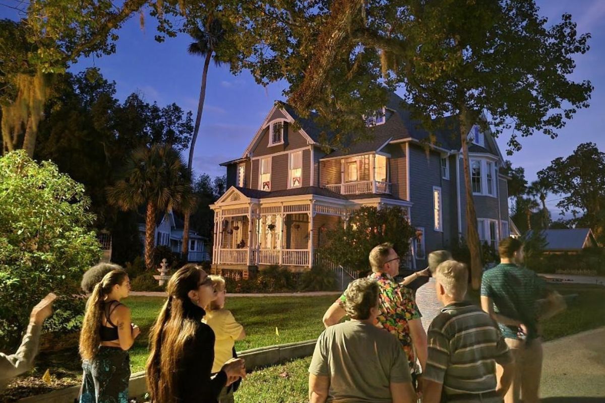 A group of people walk on a sidewalk toward a two-story gray house with white accents and a dark gray roof. Grass and trees surround the property with other houses in the background.