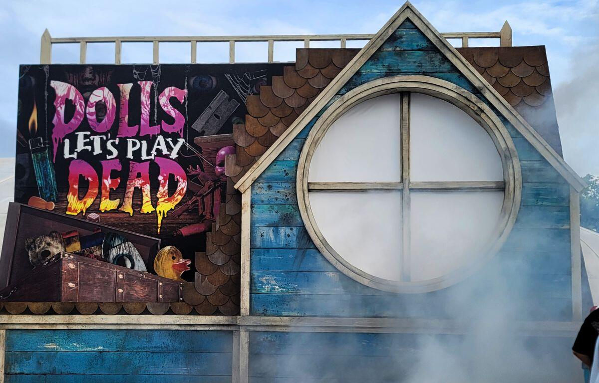 A large dollhouse facade reads, "Dolls Let's Play Dead" with scuffed toys. A razor blade and lighter surround them for a haunting look. Fog surrounds the facade.