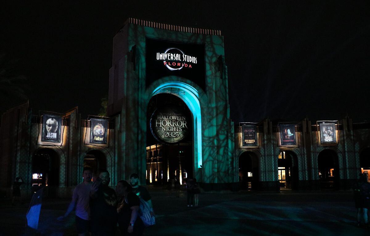 A large arch has a sign toward the top reading, "Universal Studios Florida" with a sign hanging from the middle of the arch reading, "Halloween Horror Nights 2025." To either side are small arches featuring the intellectual property houses featured at the event that recreate movie, show or game moments. Green and blue lighting provide an eerie atmosphere to the dark scene.