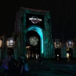 A large arch has a sign toward the top reading, "Universal Studios Florida" with a sign hanging from the middle of the arch reading, "Halloween Horror Nights 2025." To either side are small arches featuring the intellectual property houses featured at the event that recreate movie, show or game moments. Green and blue lighting provide an eerie atmosphere to the dark scene.