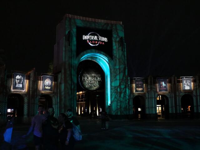 A large arch has a sign toward the top reading, "Universal Studios Florida" with a sign hanging from the middle of the arch reading, "Halloween Horror Nights 2025." To either side are small arches featuring the intellectual property houses featured at the event that recreate movie, show or game moments. Green and blue lighting provide an eerie atmosphere to the dark scene.