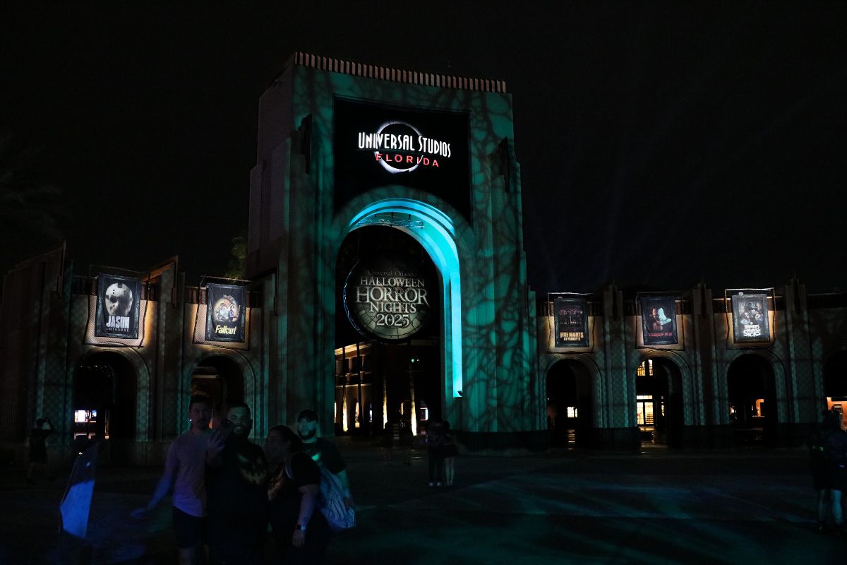 A large arch has a sign toward the top reading, "Universal Studios Florida" with a sign hanging from the middle of the arch reading, "Halloween Horror Nights 2025." To either side are small arches featuring the intellectual property houses featured at the event that recreate movie, show or game moments. Green and blue lighting provide an eerie atmosphere to the dark scene.