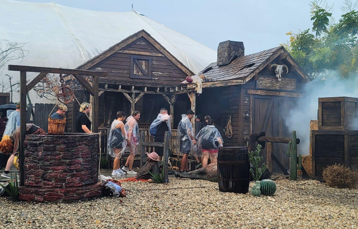 A wooden house facade decorated to look worn down and western has fake bodies strewn around. Fog fills the right side of the house and a large, white tent stands behind the house. Guests wearing ponchos enter the house in a single file line.