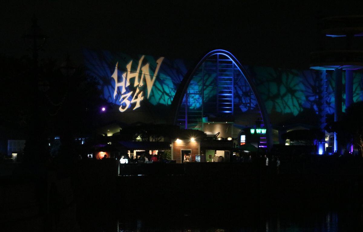 A large dark building is in the distance with a large projection of "HHN 34" on the front of the building. Green and blue lights illuminate the building creating an eerie atmosphere.