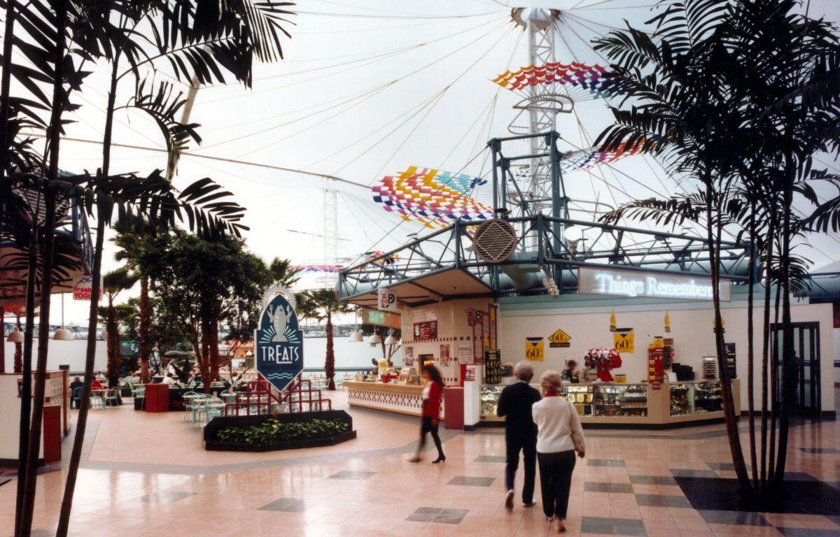 People walking around an enclosed area in a mall, with a sign that says, "food court," covered by a white canopy.
