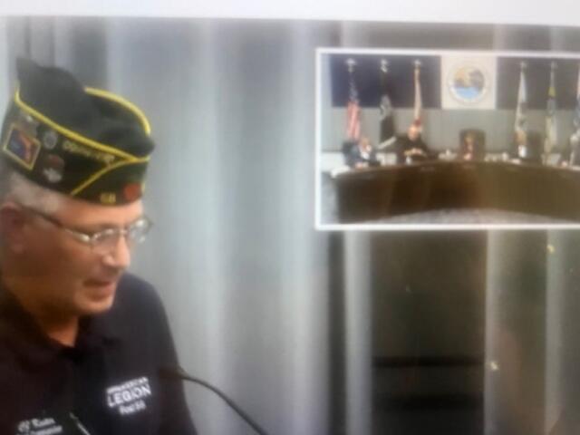 A man wearing a hat, with gray and white in the background. The man with the hat is standing at a podium with a microphone. To the right is a picture inset with people sitting behind a dais.