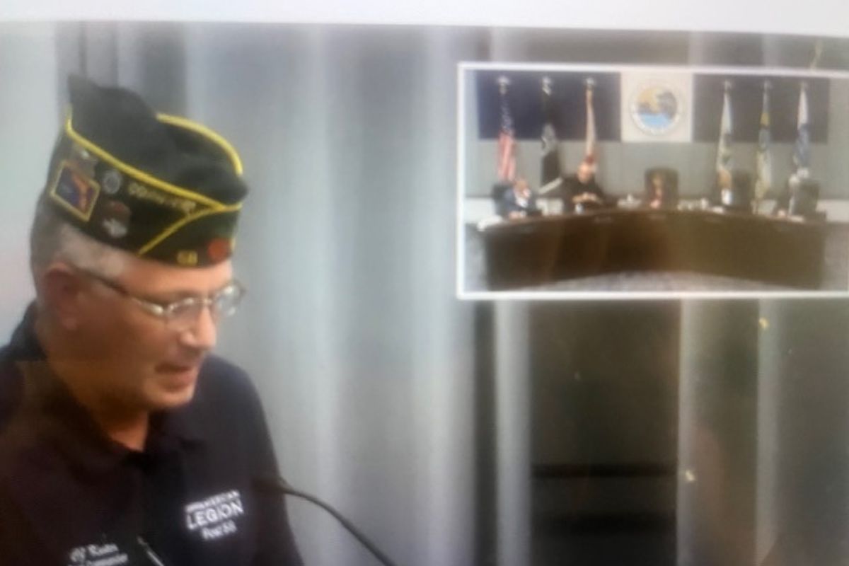 A man wearing a hat, with gray and white in the background. The man with the hat is standing at a podium with a microphone. To the right is a picture inset with people sitting behind a dais.