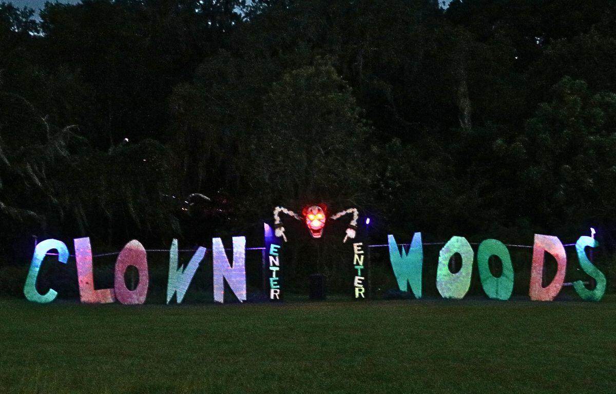 A large display of letters with different colors reads, "CLOWN WOODS" in a horizontal line and two colorful "ENTER" signs in a vertical line in the center. Between the two vertical signs is an entrance to a dark forest with a clown pointing down above the entrance.