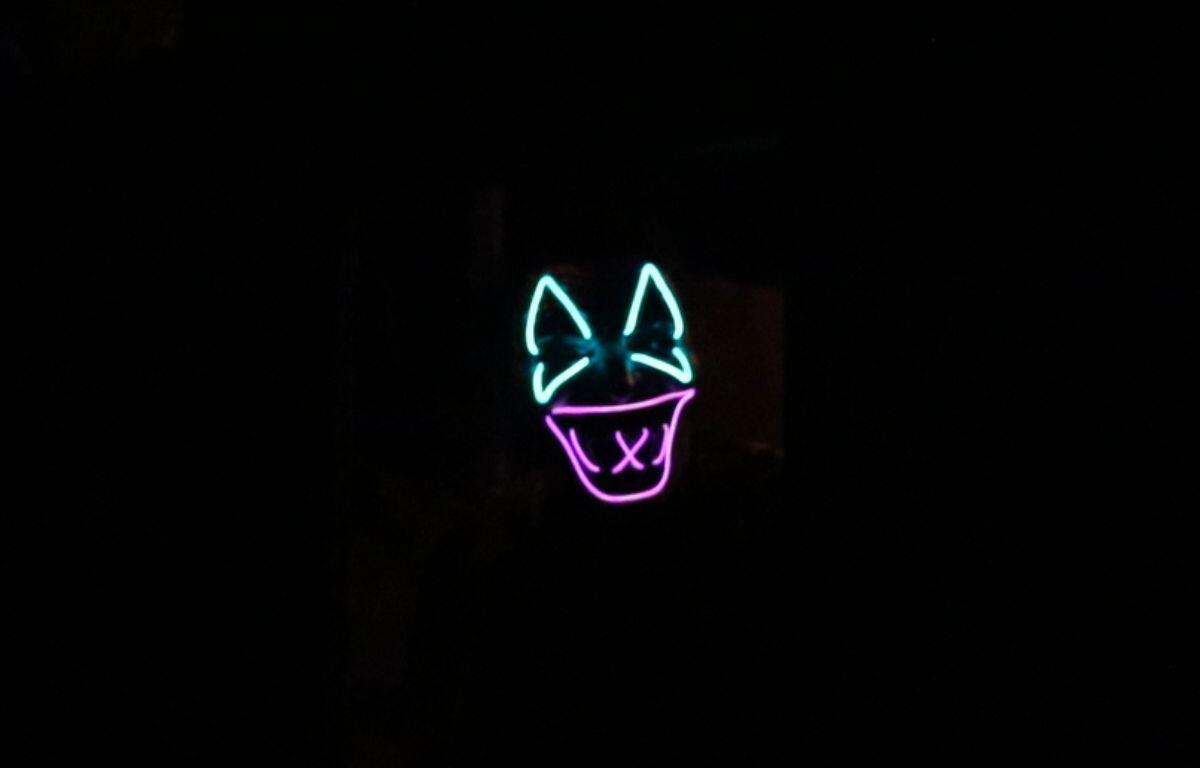 A neon green and magenta light-up mask emerges from a dark area. Eyes are barely visible beyond the green section.