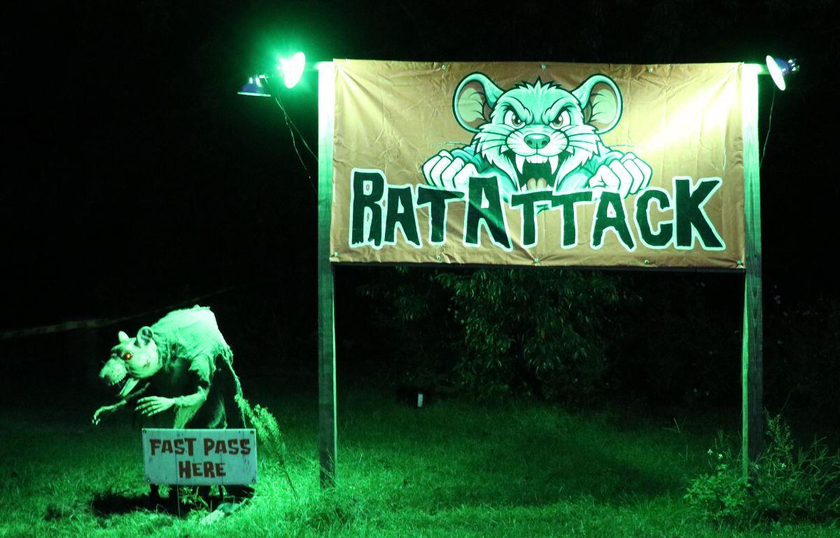 An orange sign with an angry rat and black words reading, "Rat Attack" stands in a grassy field with a dark forest behind it. An animatronic rat stands to the left of it with a white sign reading "fast pass here." Two green lights illuminate the sign and animatronic rat.