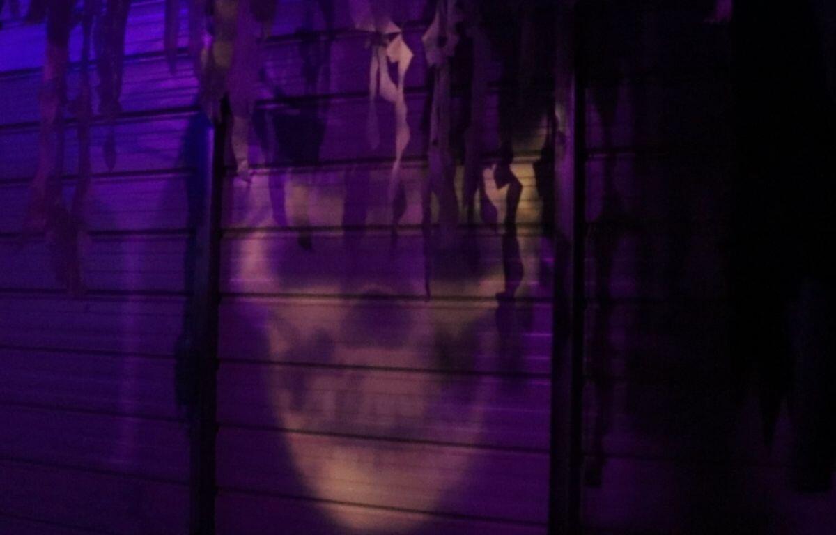 A yellow skull is projected onto a wall of wood planks with cloth hanging from the rafters.