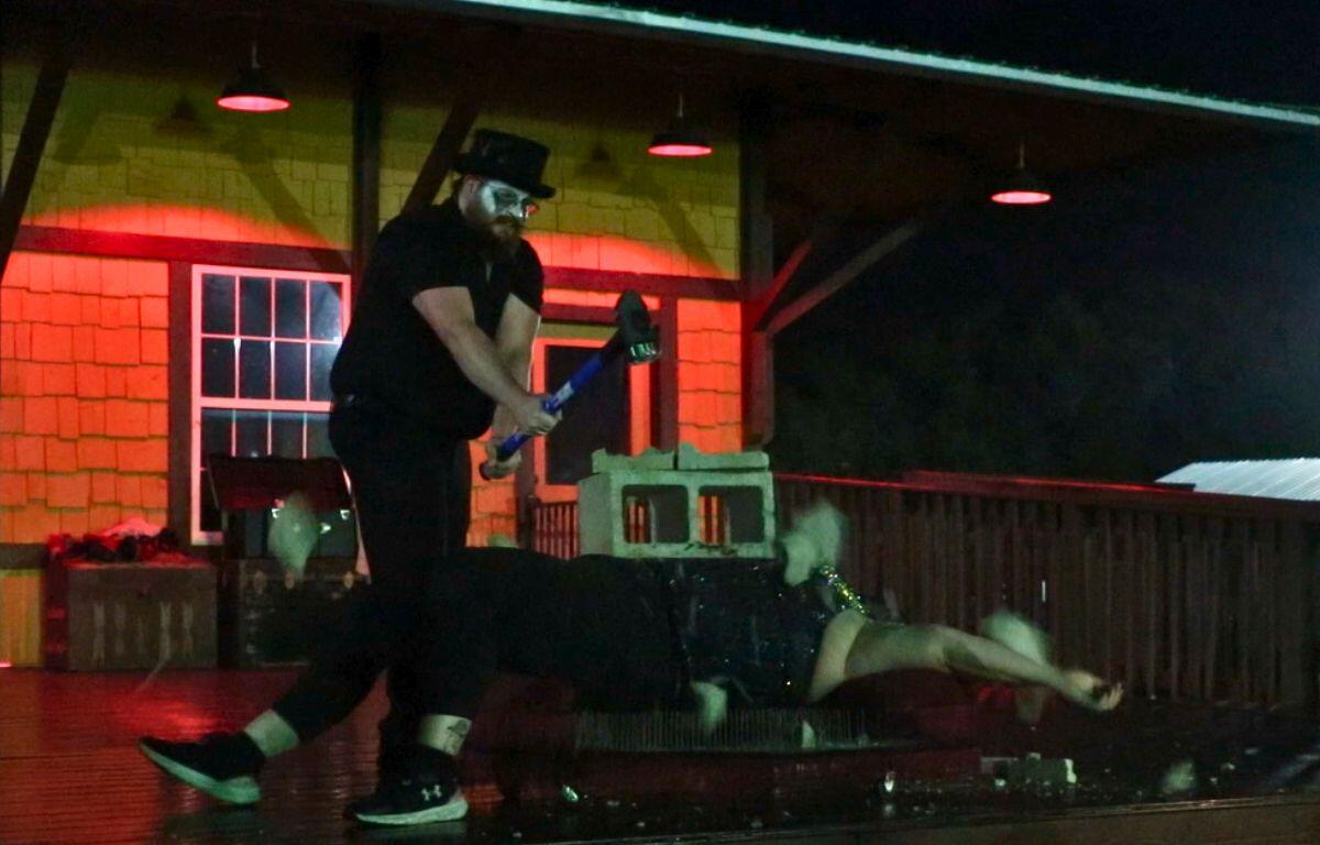 A woman lays on a bed of nails with cinder blocks on top of her. Nearby a man is swinging a sledgehammer upward. Pieces of the cinder block are flying out off of a stage. In the background is a train depot with red lighting.