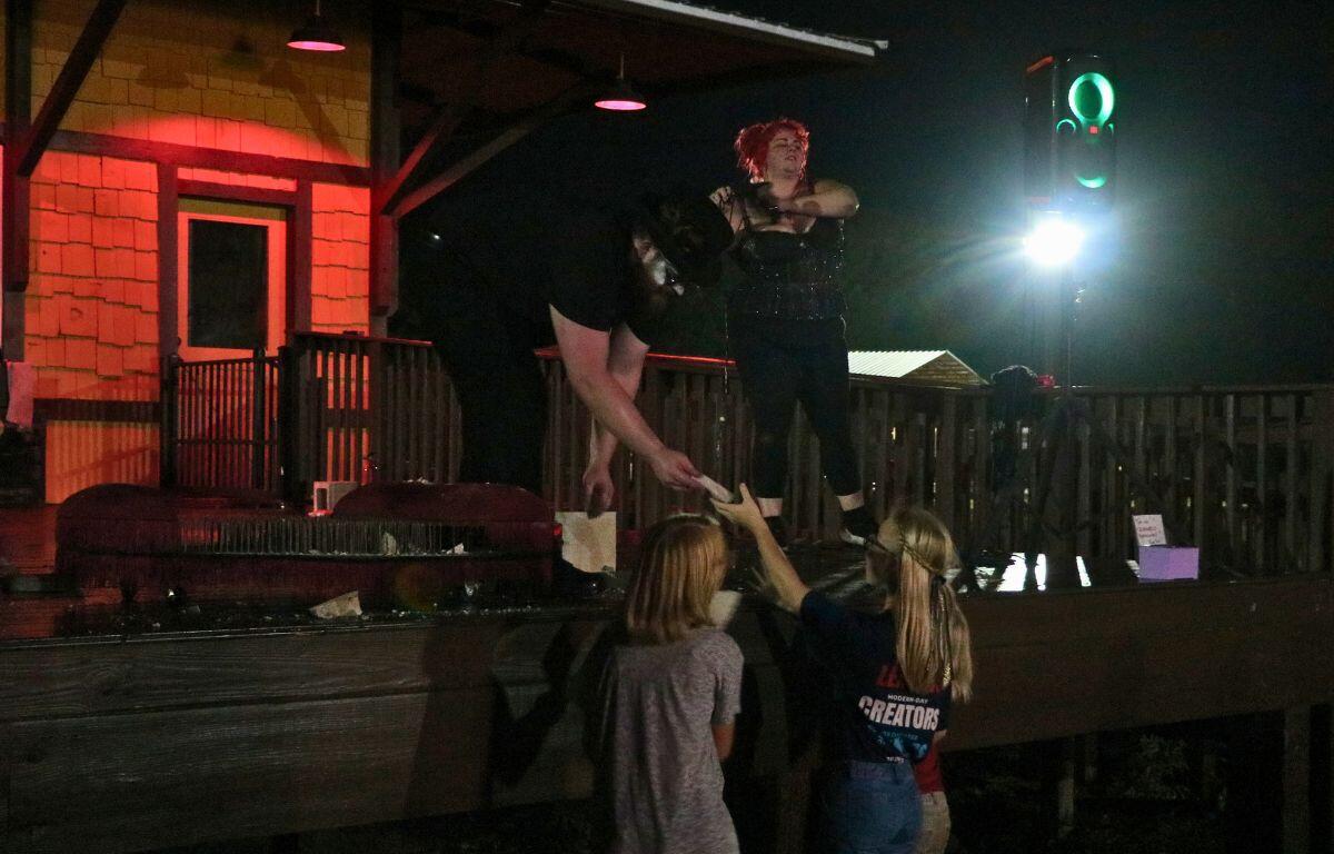 Two stage performers hand out concrete pieces to kids from a stage. Next to one of the performers is a bed of nails in the shape of a heart. In the background is a train depot.
