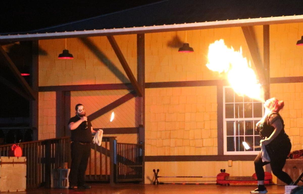 Two stage performers hold open flames in front of a train depot while the female performer blows fire with alcohol.