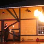 Two stage performers hold open flames in front of a train depot while the female performer blows fire with alcohol.