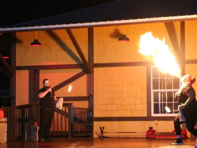Two stage performers hold open flames in front of a train depot while the female performer blows fire with alcohol.