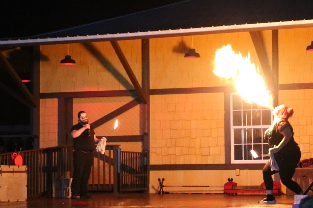Two stage performers hold open flames in front of a train depot while the female performer blows fire with alcohol.