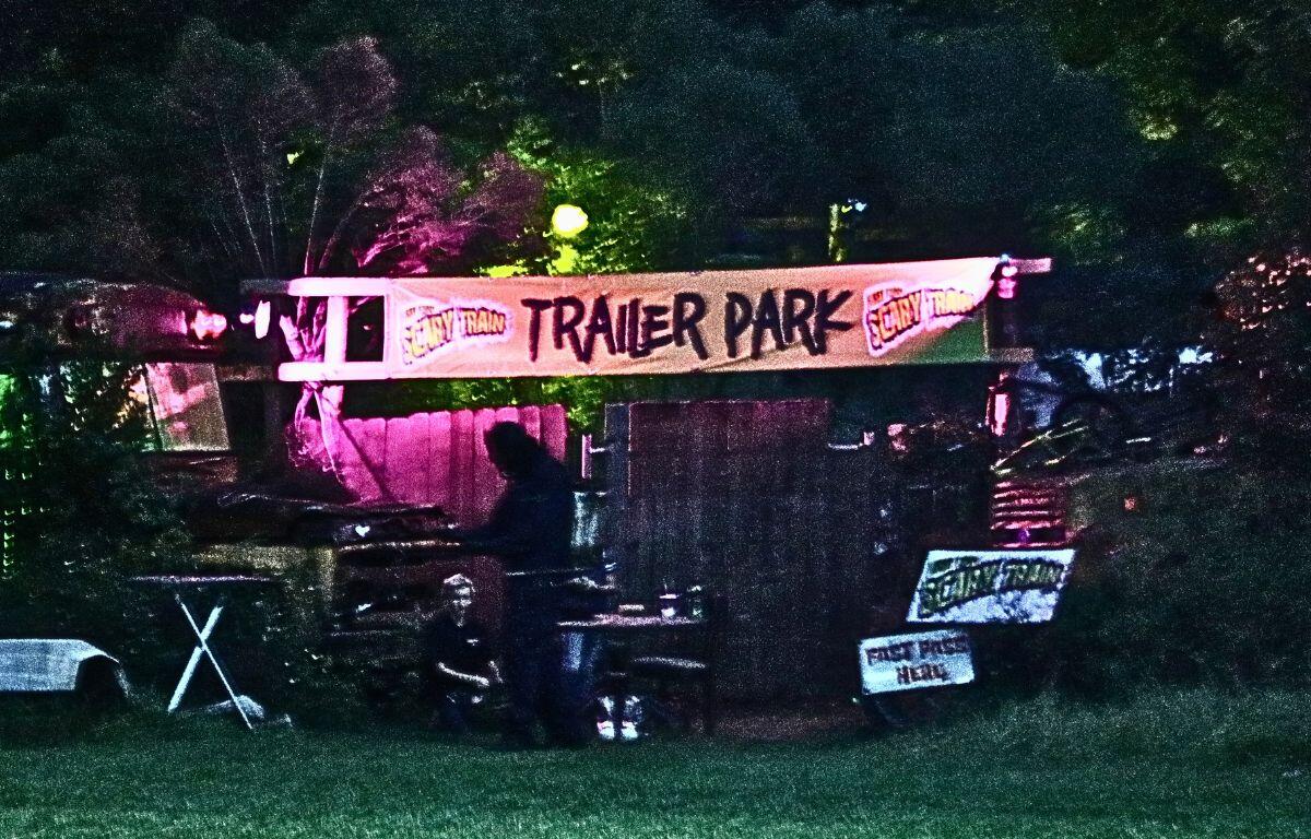 A wood wall, broken cars and two kids block the entrance of a trailer park haunted attraction. In the background is a forest with minimal lighting. In the front is a sign reading, "Scary Train Trailer Park."