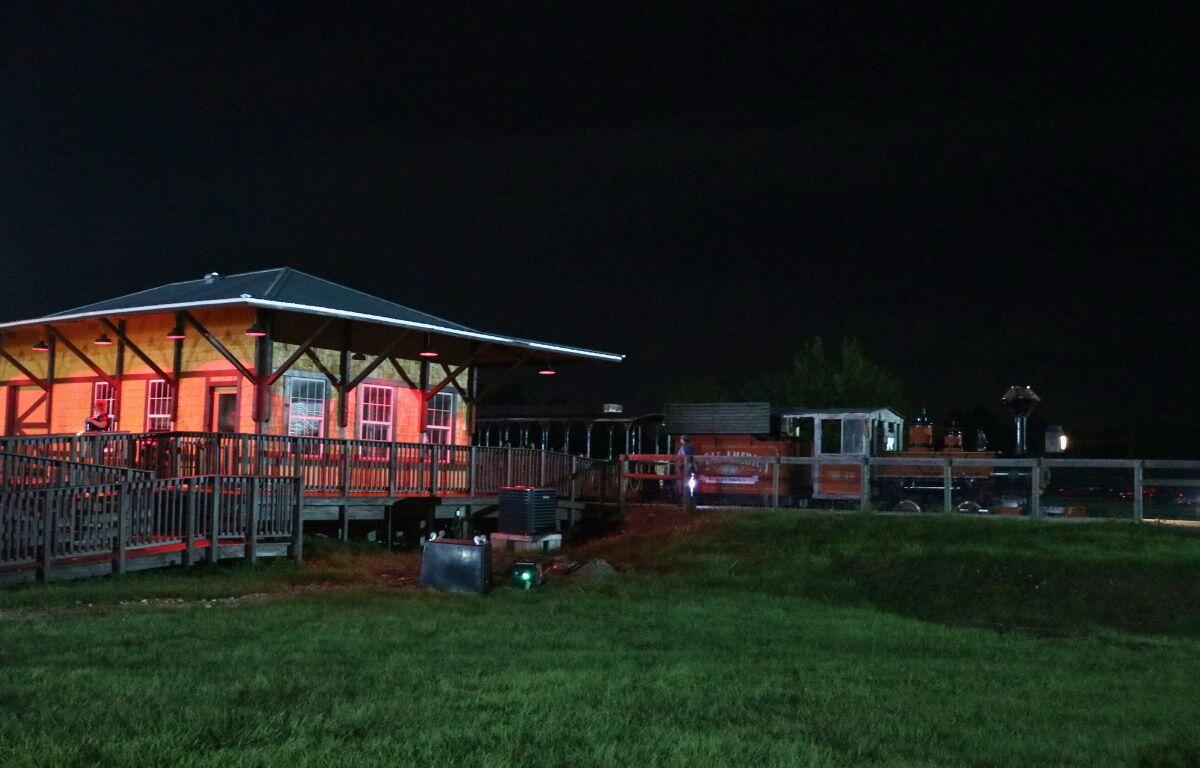 A train depot with red lighting stands in an open field toward the left while a train begins to leave the station toward the right. In the background is a dark forest.