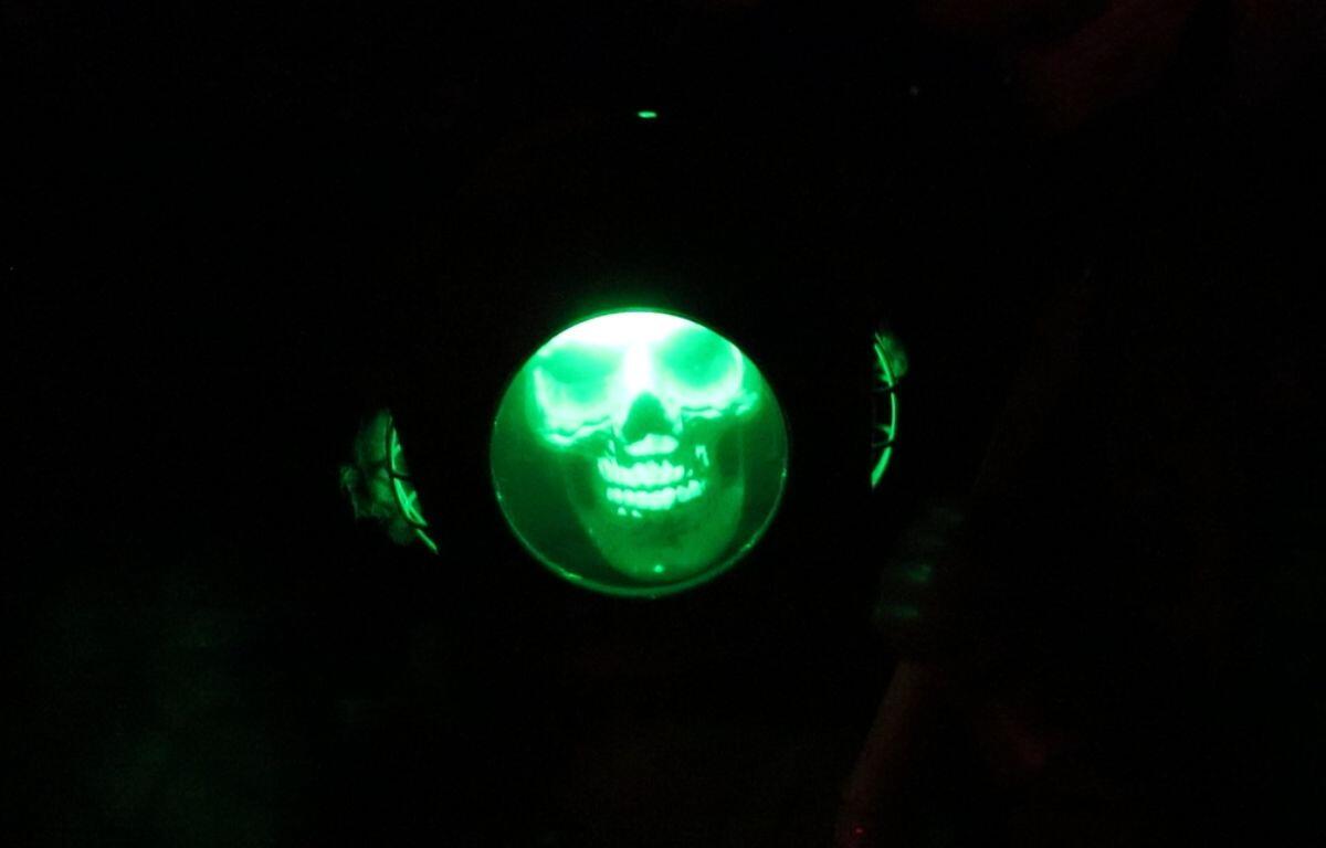 A skeleton in a standard driving dress stands alone in a dark room with a green light illuminating it from inside of the helmet.