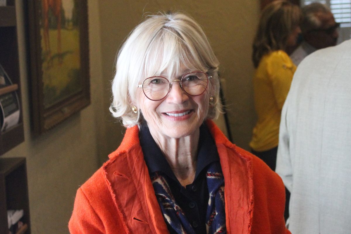 A woman wearing a red sweater, standing in front of a room with people, with art against the left wall.