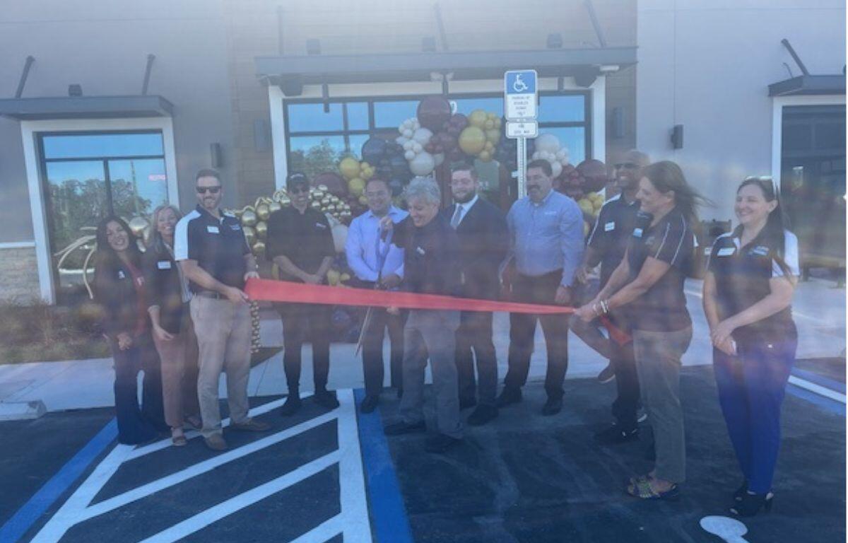 In the foreground a paved lot with lines. In the middle of the photo, a group of people standing and holding a red ribbon, with the man in the middle holding an oversized pair of scissors. in the background, the area is festooned with ballons, in and there's a parking sign, in front of t building with windows.
