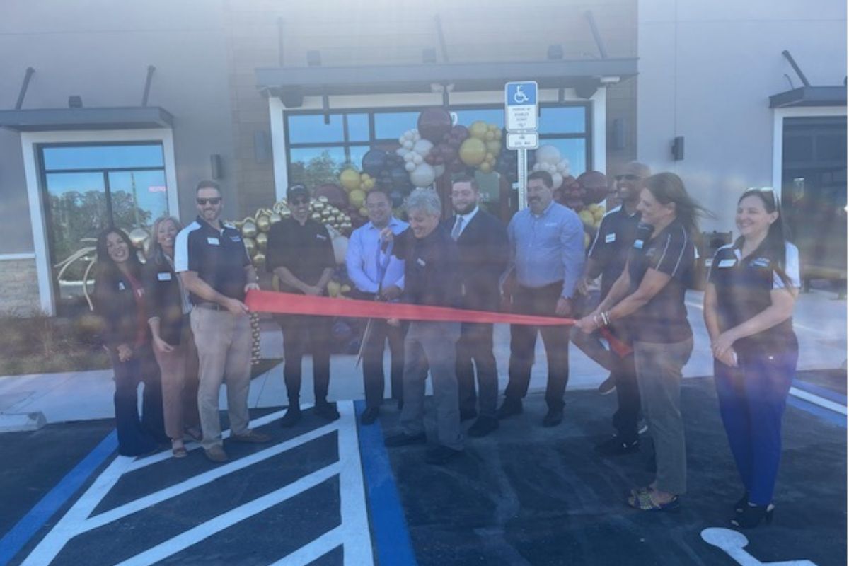 In the foreground a paved lot with lines. In the middle of the photo, a group of people standing and holding a red ribbon, with the man in the middle holding an oversized pair of scissors. in the background, the area is festooned with ballons, in and there's a parking sign, in front of t building with windows.