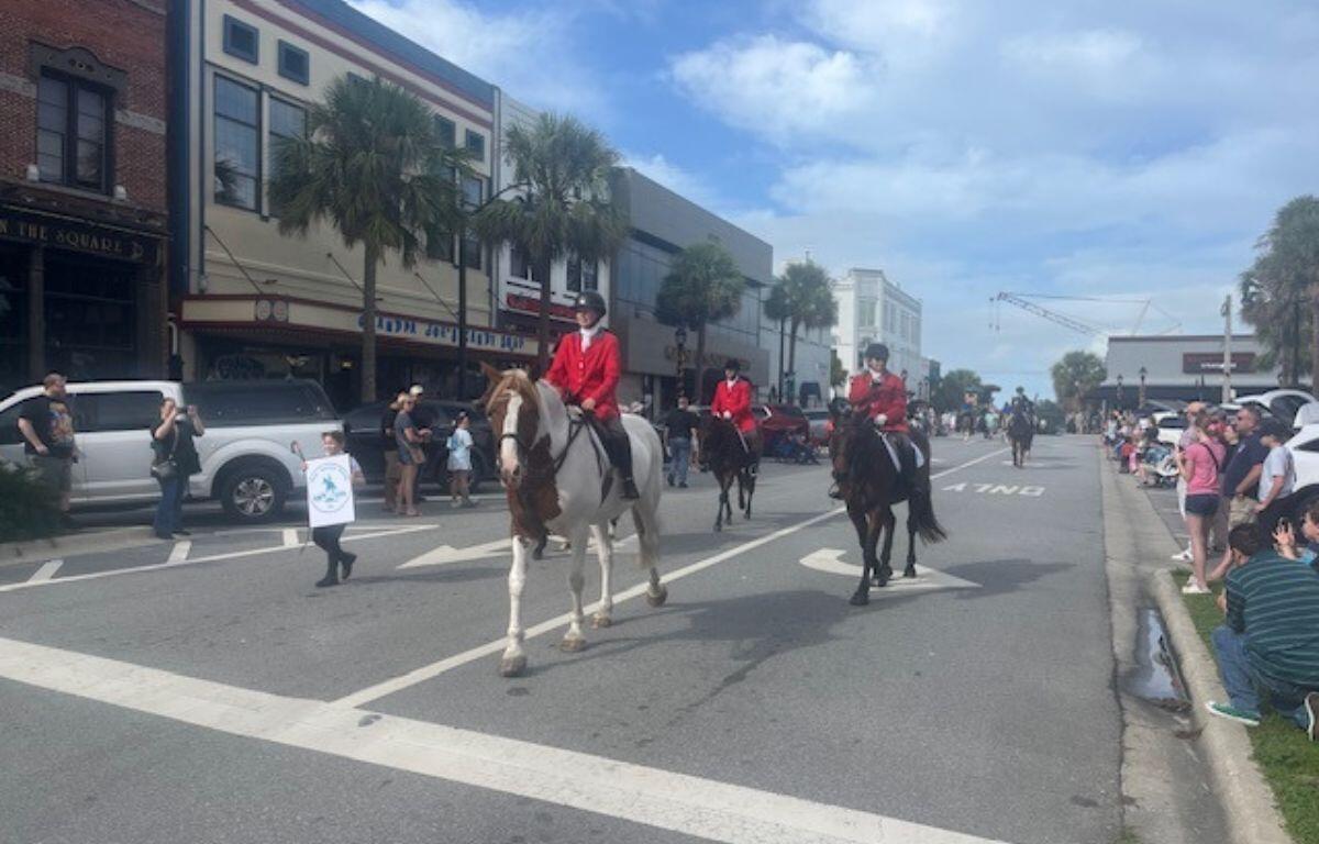 On the left, store fronts, palms and a person holding a placard, in the center horses with riders in formal riding attire and on the right the thoroughfare and people lining the street, and lines on the street foreground.