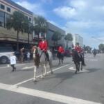 On the left, store fronts, palms and a person holding a placard, in the center horses with riders in formal riding attire and on the right the thoroughfare and people lining the street, and lines on the street foreground.