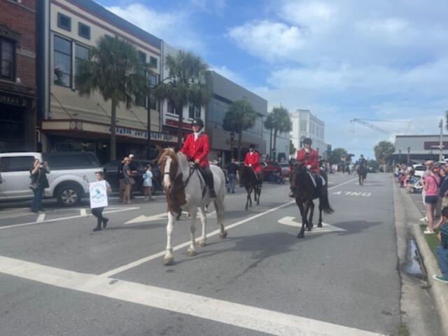 On the left, store fronts, palms and a person holding a placard, in the center horses with riders in formal riding attire and on the right the thoroughfare and people lining the street, and lines on the street foreground.