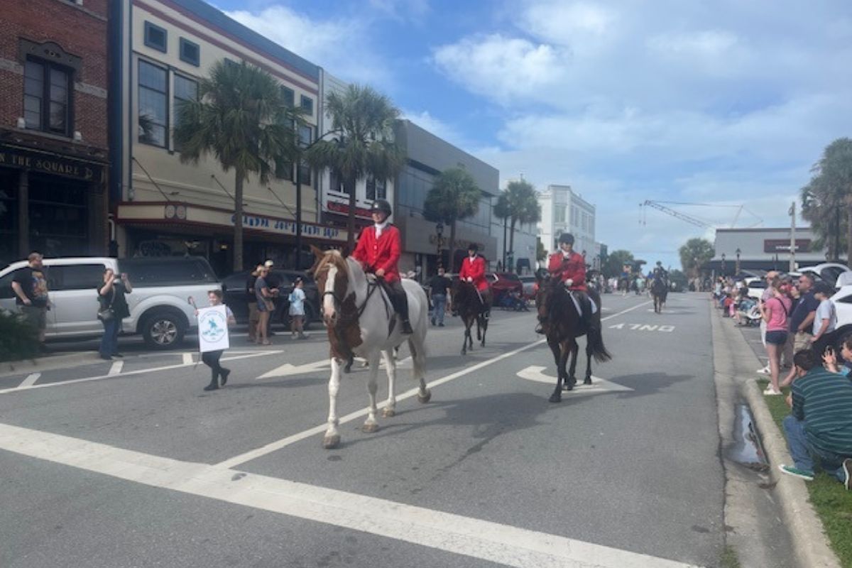 On the left, store fronts, palms and a person holding a placard, in the center horses with riders in formal riding attire and on the right the thoroughfare and people lining the street, and lines on the street foreground.