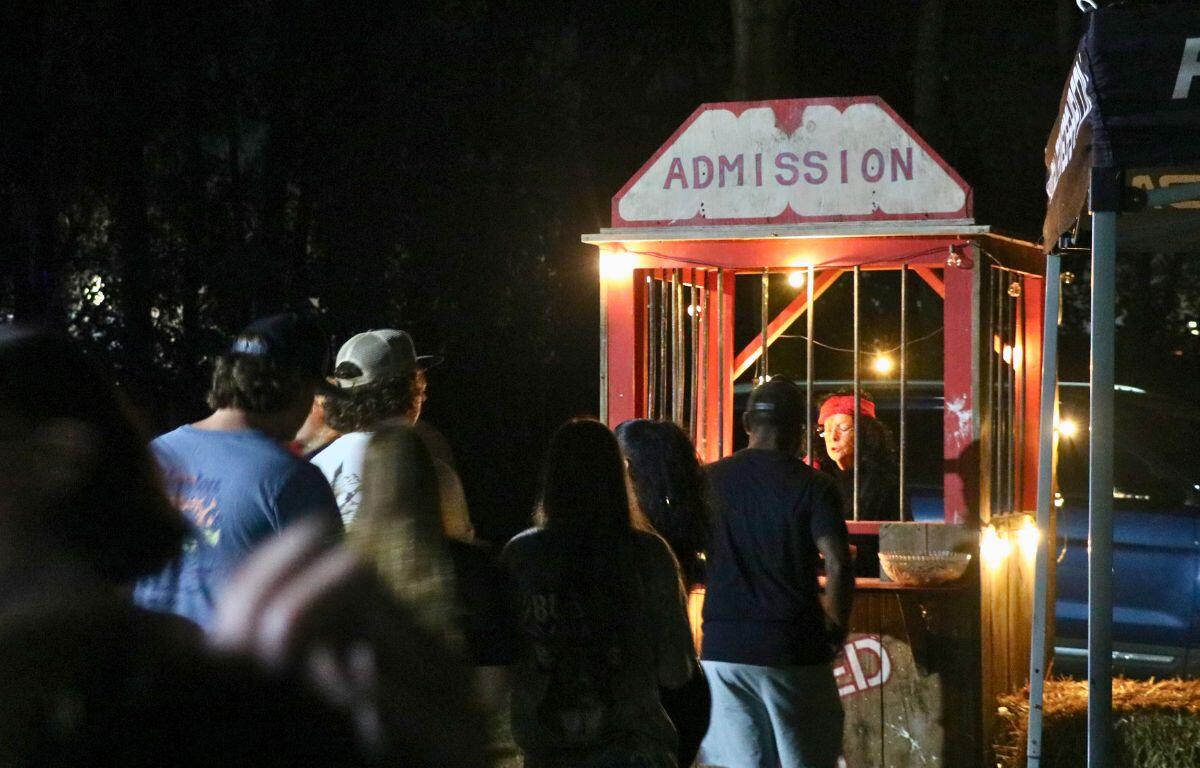 A wooden red and white ticket booth with "Admission" written at the top and a woman in a costume stands at the end of a line queue. In the background is a dark and dense forest.