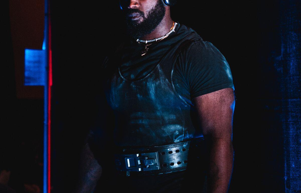 A man standing against a dark background, wearing a dark t-shirt, weightlifting belt and doo-rag.