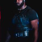 A man standing against a dark background, wearing a dark t-shirt, weightlifting belt and doo-rag.