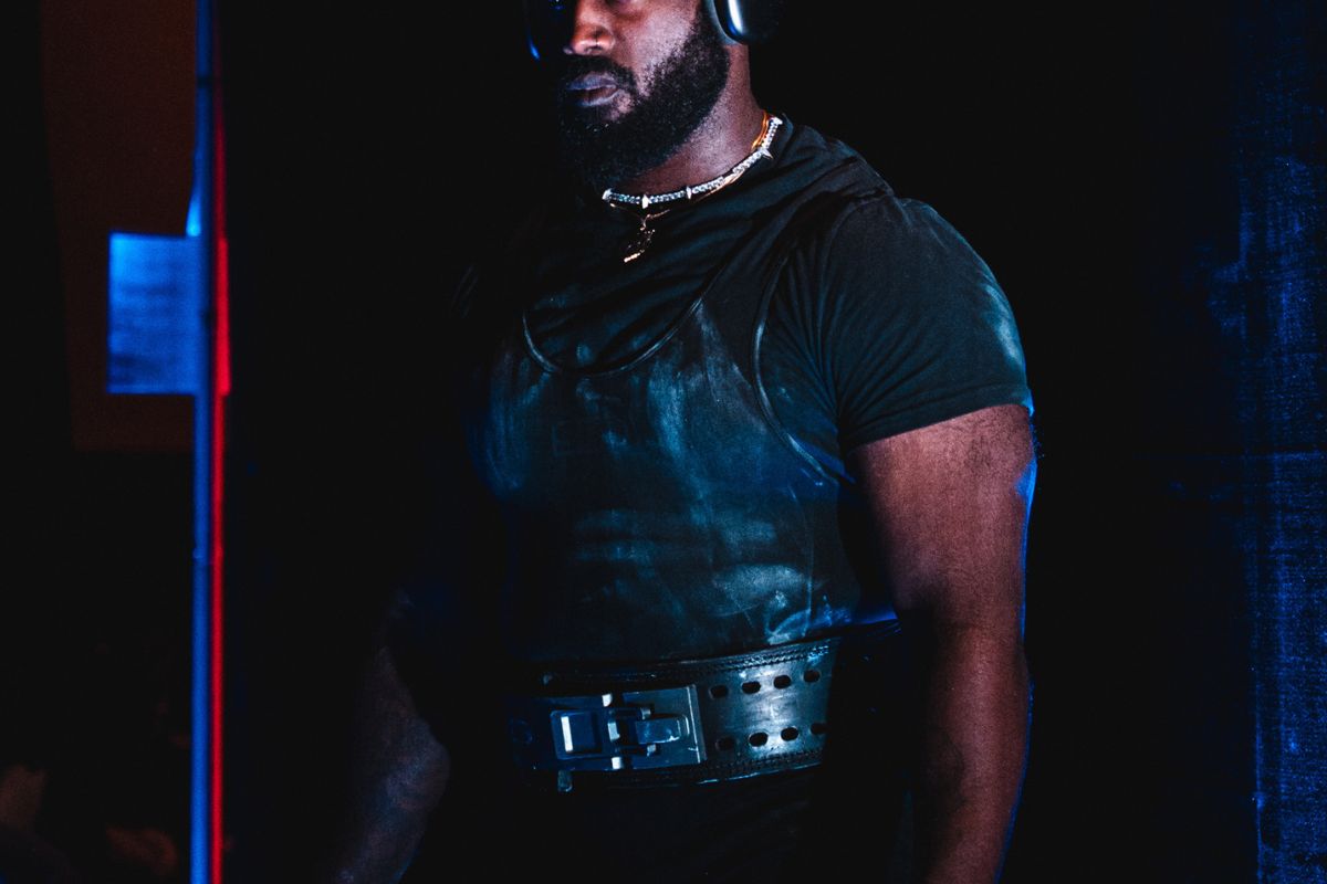 A man standing against a dark background, wearing a dark t-shirt, weightlifting belt and doo-rag.