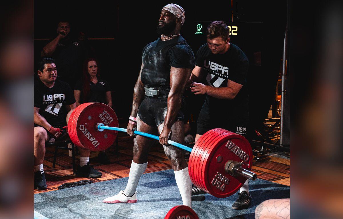 A man lifting a bar with weights on both the left side of the bar, standing on a mat that's also in the foreground, and a man standing behind him, spotting him, with people to the left, center and right in the background.